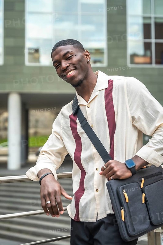 Young black man smiling and posing with laptop bag on his shoulder, leaning against a railing outside a modern building, representing urban lifestyle and professional confidence