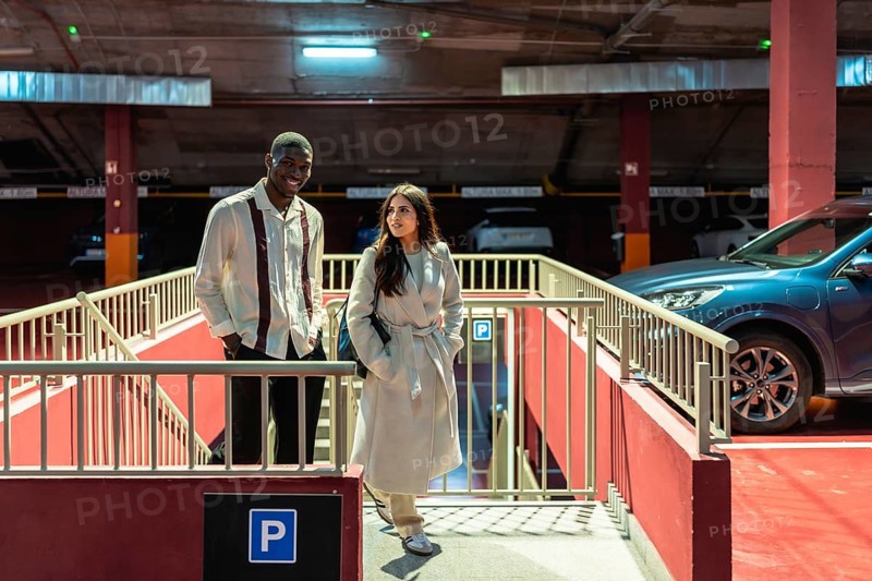 Diverse interracial couple walking up concrete stairs in an urban parking garage, smiling and holding hands as they exit underground, enjoying a casual city outing together