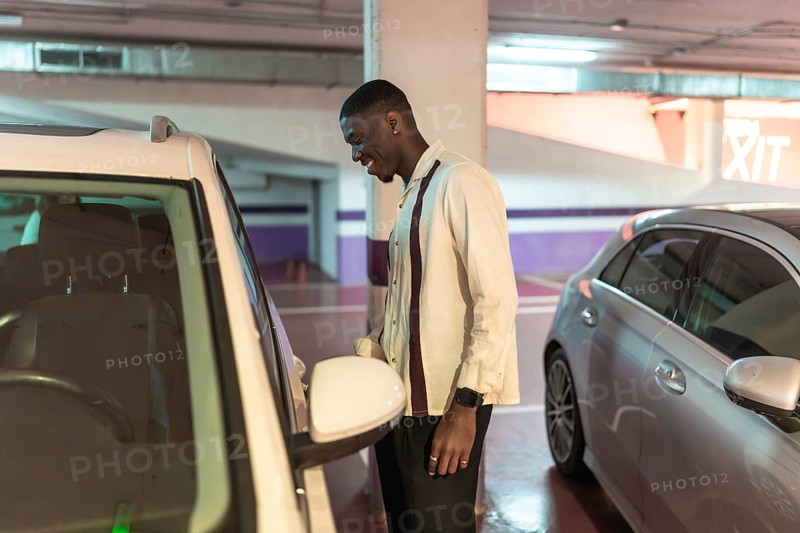 Young black man in an underground parking garage smiling as he opens his white car door, confident and ready for a nighttime urban drive, travel or arrival into the city