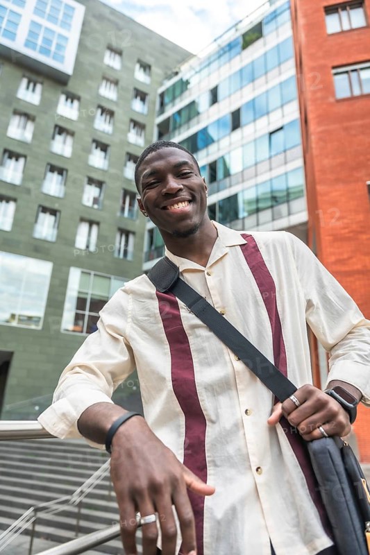 Young black man smiling and leaning on a railing in a modern city street, carrying a bag and dressed business casual, representing confident urban student and young professional life