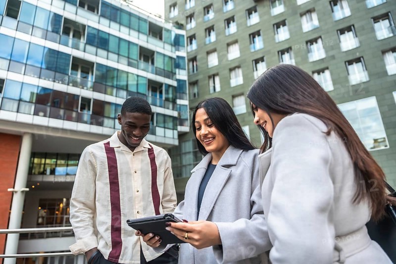 Diverse business professionals smiling and collaborating while reviewing data together on a touchscreen tablet outside an office building, demonstrating teamwork and modern communication