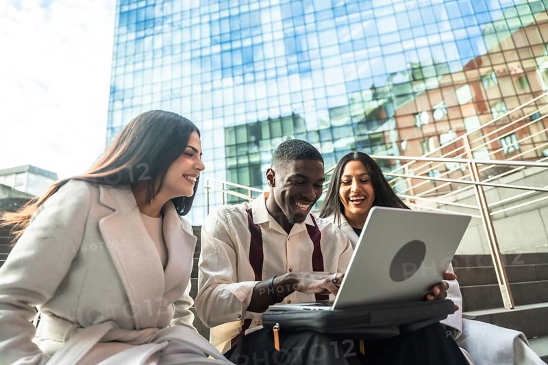 Group of diverse coworkers laughing while sharing content and collaborating on a laptop during an outdoor meeting in an urban setting, reflecting teamwork and success