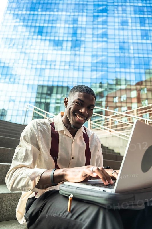 Young black man smiling while typing on a laptop, sitting on urban stairs with a modern glass building reflecting the blue sky in the background, working or studying