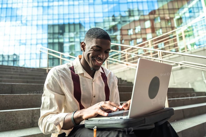 Young black man sitting on concrete stairs in a city, smiling and typing on a laptop, portraying remote work, modern technology, urban lifestyle and professional focus outdoors