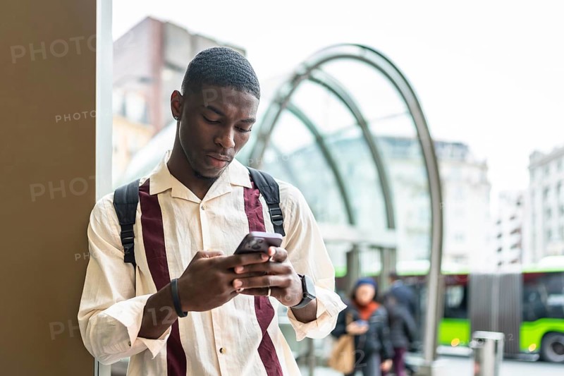 Young african american man with backpack focused on smartphone while waiting at a city bus stop, commuting and browsing apps in a modern urban downtown setting