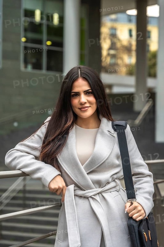Businesswoman posing with confidence, holding her laptop bag and leaning on a railing, portraying professionalism and success in a modern urban setting