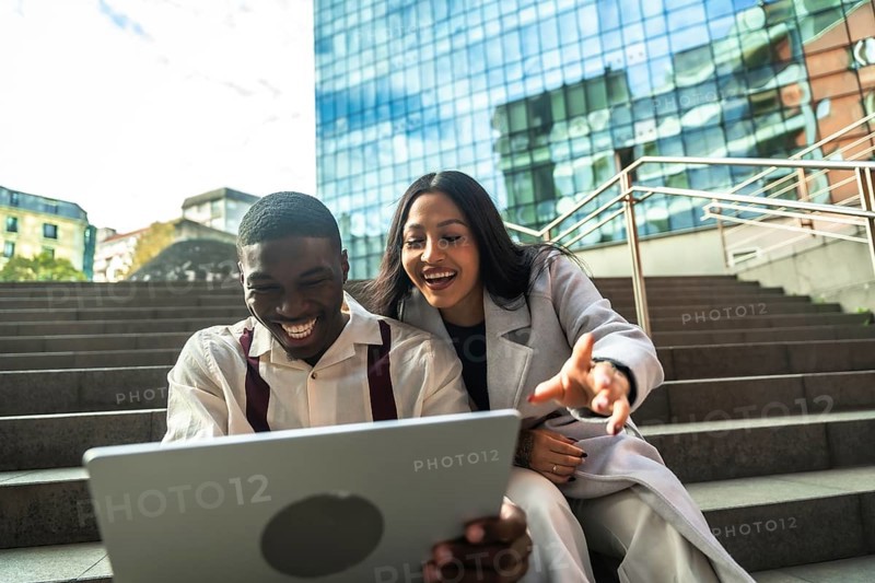 Diverse business colleagues sitting on steps, smiling and laughing while looking at a laptop together, enjoying a break outside a modern office building in the city