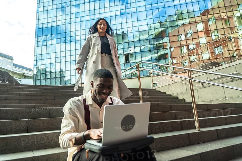 Young professional smiling at laptop on urban steps while a stylish woman walks down nearby by a modern glass building, capturing outdoor remote work and city life ambiance