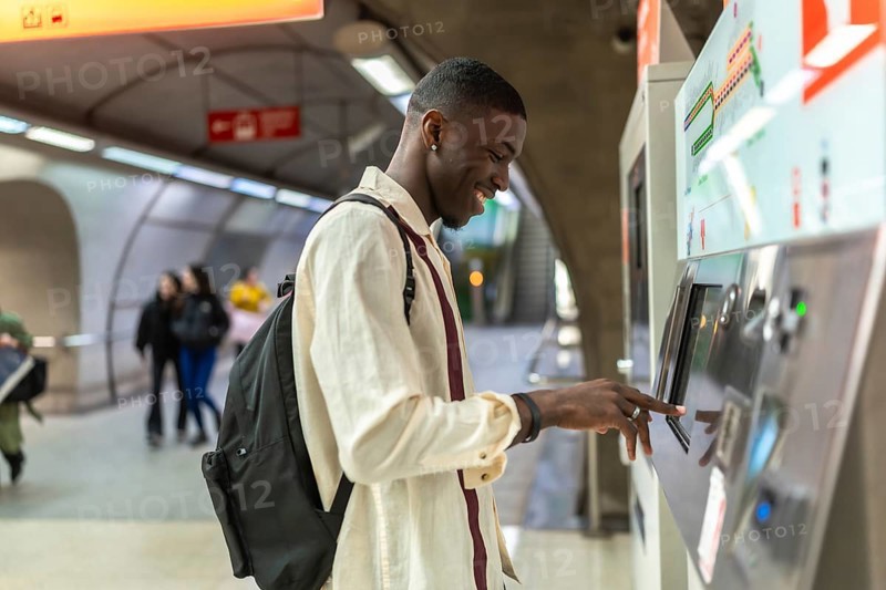Young man standing at an automated ticket machine, smiling, and touching the screen while purchasing public transport fare in a modern urban subway station
