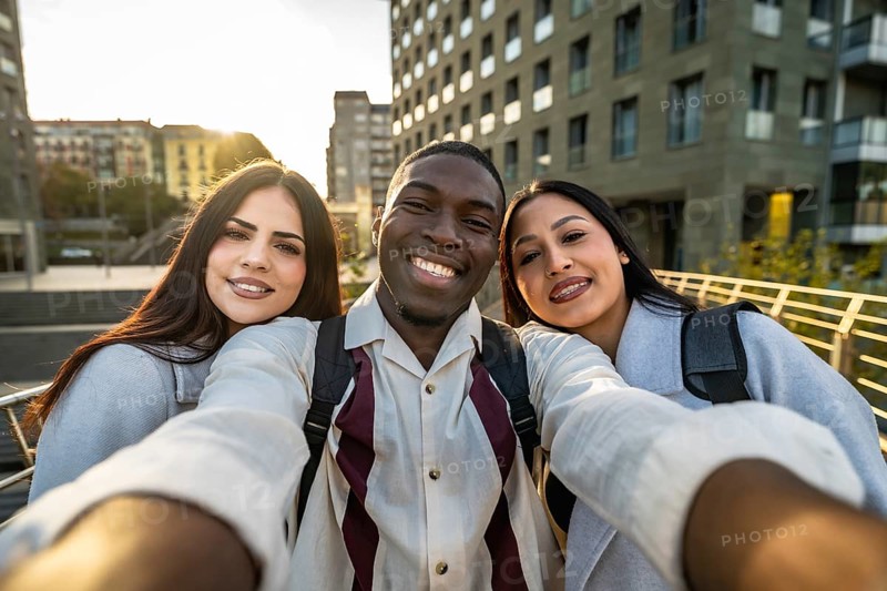 Three diverse students smiling into a smartphone, taking a city street selfie while walking and celebrating friendship, youth, and multicultural college life in warm daylight