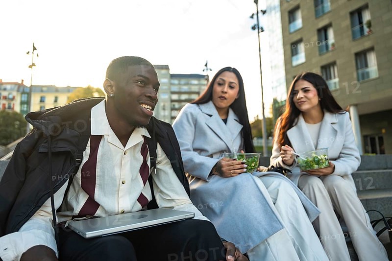 Diverse group of young adult colleagues or friends smiling and having a relaxed conversation while eating healthy salads on stairs during their outdoor lunch break in a city setting