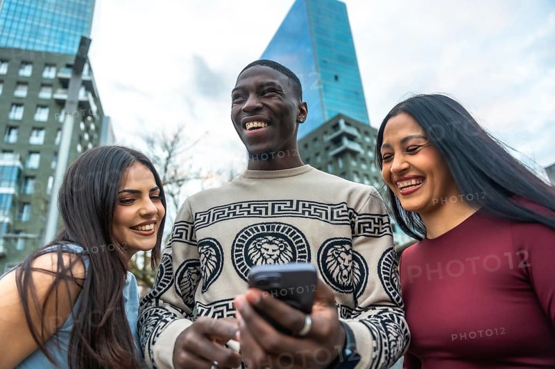 Diverse group of young adult friends laughing, smiling, and happily sharing content on a smartphone outdoors in an urban environment with modern buildings