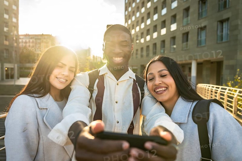 Group of diverse young adults smiling and having fun together while taking a selfie with a smartphone, capturing happy moments in an urban setting with golden hour light