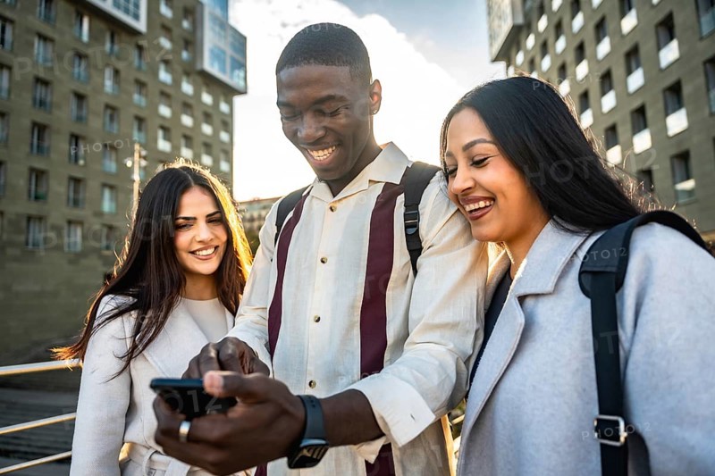 Diverse group of young friends laughing and sharing smartphone content outdoors in a sunny urban setting, enjoying casual connection, social media browsing and togetherness