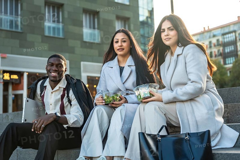 Diverse colleagues sitting on steps and smiling while enjoying healthy salads during a casual urban lunch break, representing modern work life balance and friendship