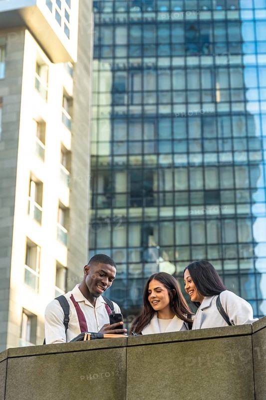 Three diverse students, a man and two women, smiling and looking at a smartphone, sharing content and interacting socially outdoors in a modern city environment