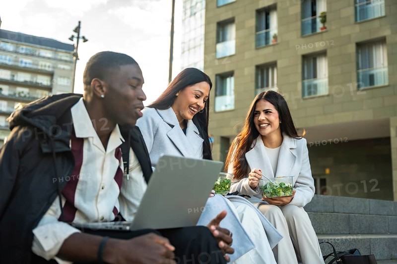 Three smiling diverse business people sitting on city stairs, discussing work and happy together during a successful outdoor lunch break with laptop and fresh salad