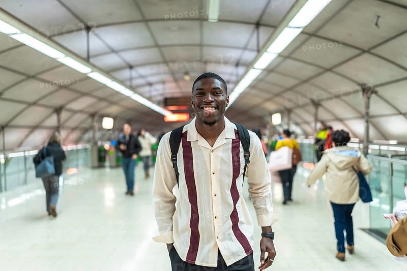 Young african man smiling, wearing a backpack and casual clothes, walking confidently through a busy modern public transport station or airport corridor
