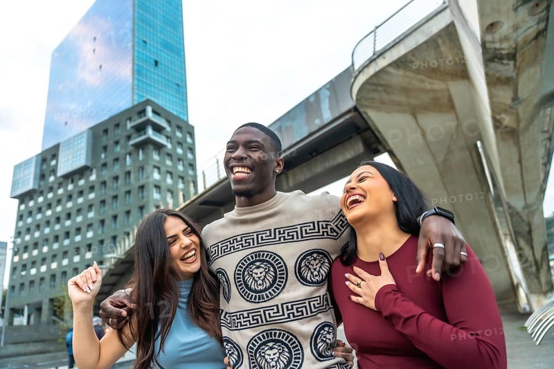 Cheerful diverse group of young adults standing closely together, smiling, laughing, and enjoying a moment of genuine friendship and happiness in a modern city environment