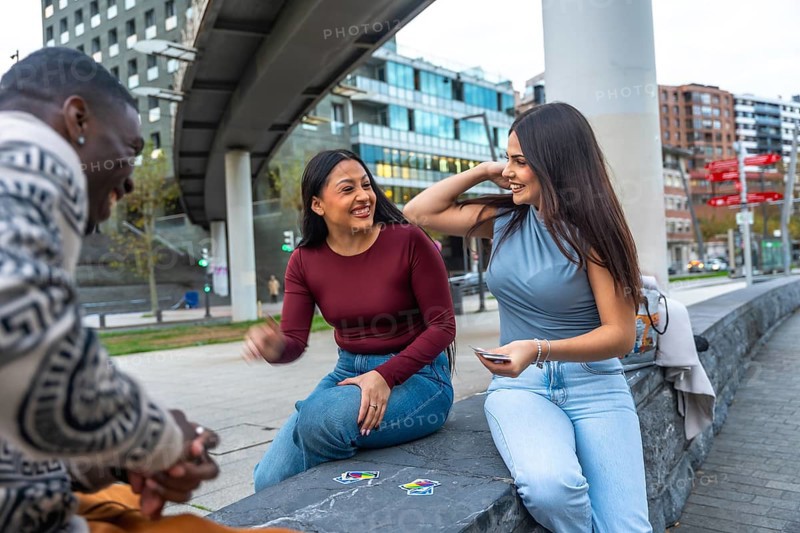 Happy diverse friends sitting in an urban park, playing cards and laughing together, enjoying relaxed outdoor leisure, conversation and casual bonding on a sunny day