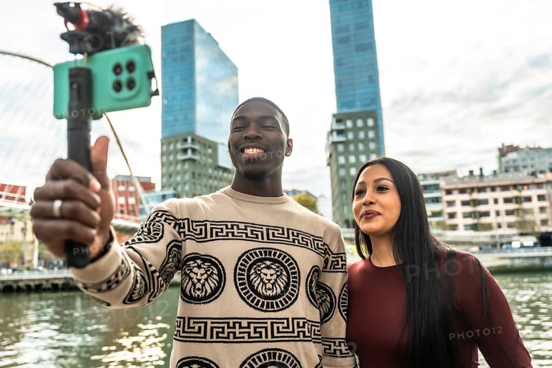 Smiling diverse couple creating content for social media, capturing a selfie video with a smartphone on a vlog stick in an urban setting with modern buildings