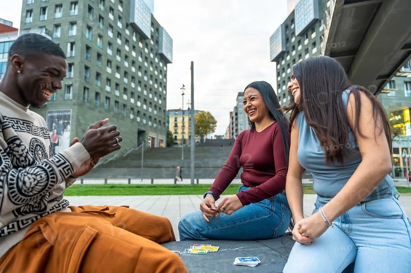 Diverse young friends sitting in a modern urban square, laughing and playing cards together outdoors, sharing a relaxed, genuine moment of friendship and leisure