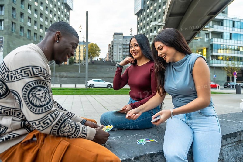 Diverse group of friends sitting outdoors in the city, smiling and laughing while playing cards, enjoying relaxed leisure time, connection and genuine youthful fun