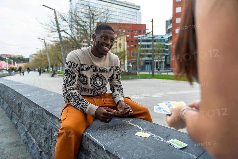 Young man smiling as he and a woman play a competitive card game on a stone bench in a modern urban park, enjoying sunny leisure, friendly competition and connection