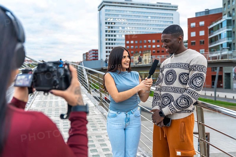 Journalist holding a microphone and interviewing a smiling young man on a modern urban bridge while a third person is filming with a professional camera