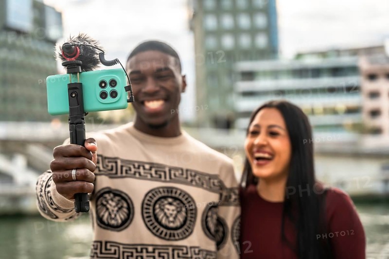 Diverse couple smiling and laughing while creating a vlog, holding a smartphone with an external microphone on a selfie stick, sharing experiences for online audiences