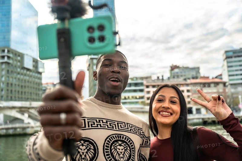 Diverse interracial couple vlogging with a smartphone on a selfie stick, smiling as they record travel content in a modern city, sharing their urban adventure and connection online