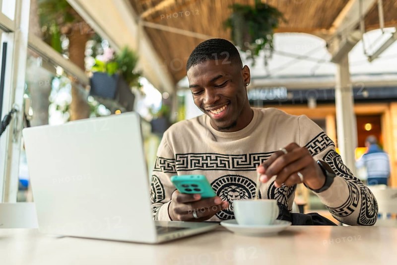 Young black man in a coffee shop using a laptop and smartphone, stirring his coffee while smiling, enjoying freelance work and digital nomad lifestyle