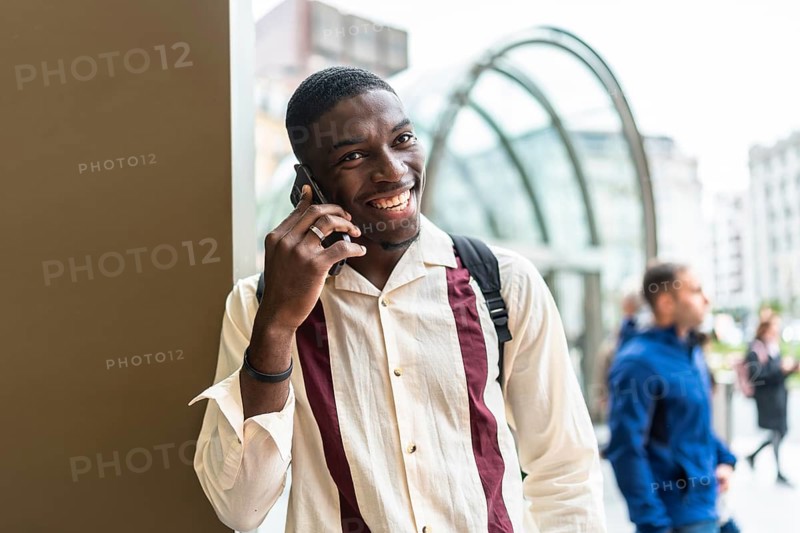 Smiling young black man talking on his mobile phone in an urban setting, enjoying a conversation outdoors in the bustling city street, highlighting modern communication and a joyful lifestyle