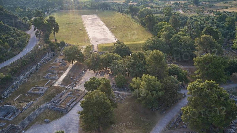 Ancient stadium in the morning backlight, ancient ruins next to a wide meadow, framed by dense tree landscape, drone shot, archaeological site, Ancient Olympia, Peloponnese, Greece