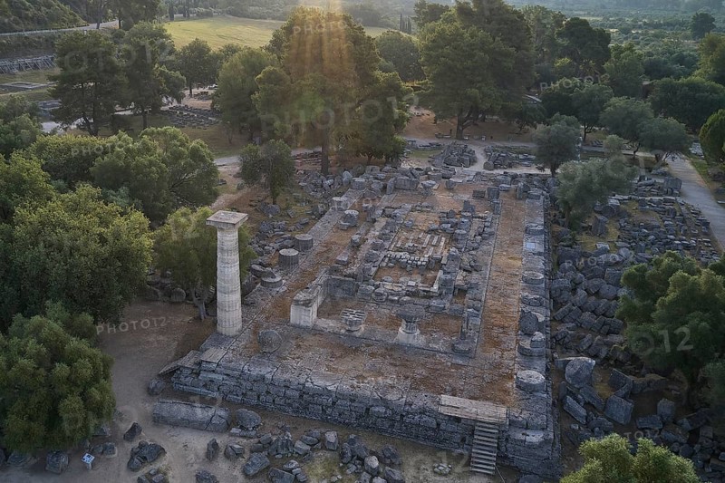Temple of Zeus in backlight, aerial view of ruins under trees with a distinctive stone pillar in sunlight, drone shot, archaeological site, Ancient Olympia, Olympia, Peloponnese, Greece