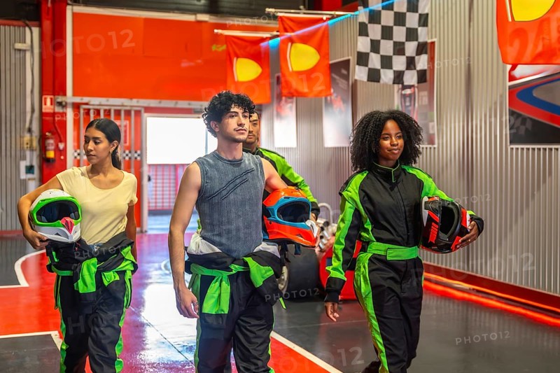 Diverse friends preparing for a go kart race, holding helmets and wearing race suits, walking through an indoor track before starting their leisure competition