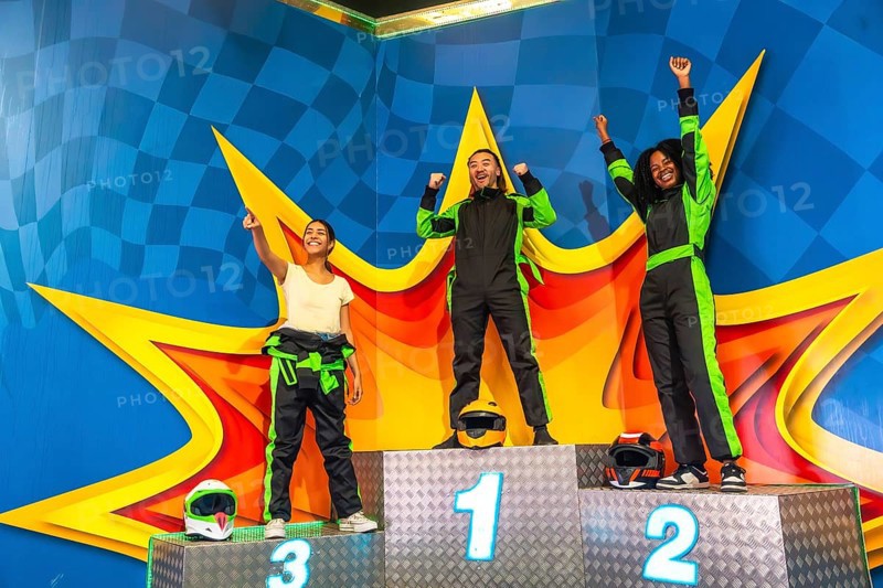 Three young multi ethnic adults celebrating their success, standing on a numbered podium wearing racing suits and helmets after a thrilling karting competition