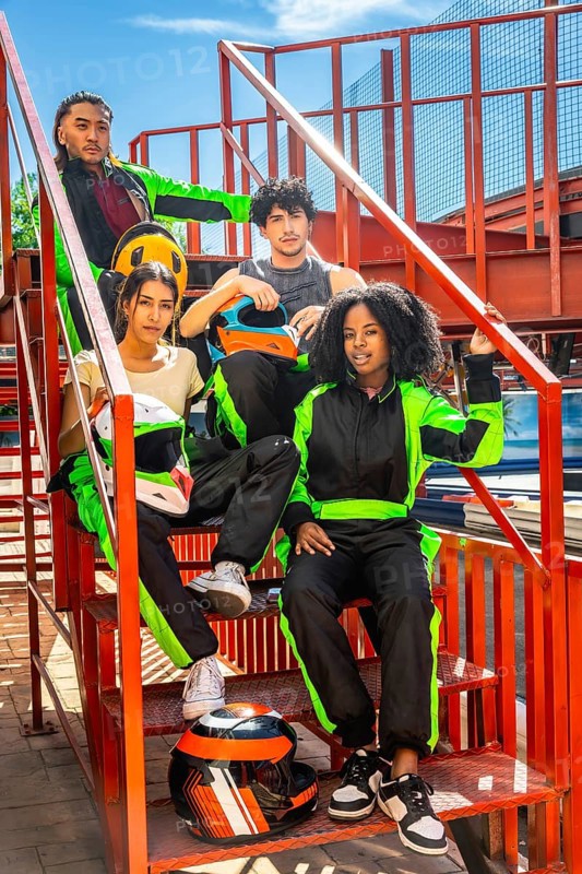 Group of diverse young people wearing racing suits and holding helmets, posing on metal stairs before starting a fun karting competition, ready for sport and leisure
