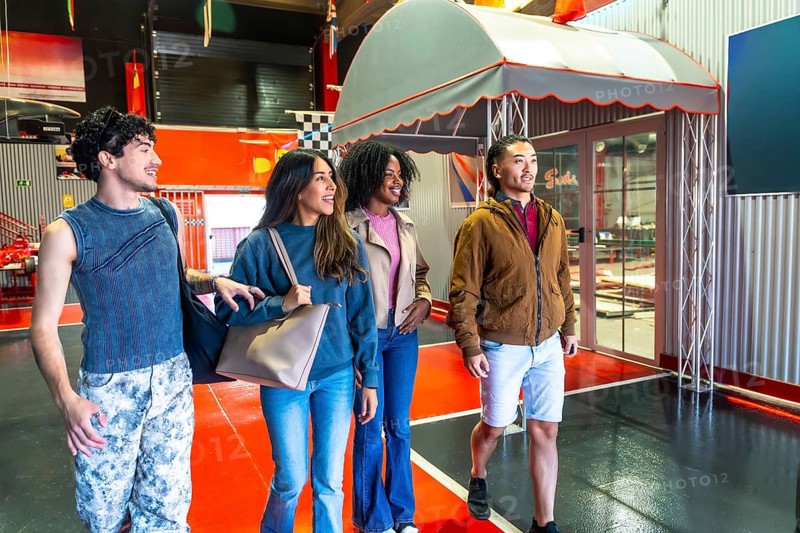 Group of diverse young adult friends smiling, walking, and looking around at an indoor karting facility, enjoying a fun leisure activity and entertainment together