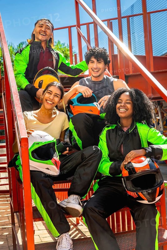 Diverse friends in racing suits holding helmets, smiling and sitting on red metallic stairs outside, celebrating the excitement and fun of karting as a group leisure activity