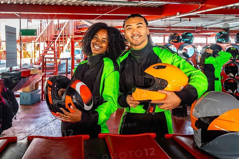 Multiracial young couple smiling at camera in racing suits and helmets, standing on indoor go kart track ready for a fun, excited karting adventure and weekend racing together