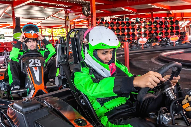 Two young men, one putting on his helmet and another holding a steering wheel, are preparing for a fun karting competition at an indoor go kart racing facility