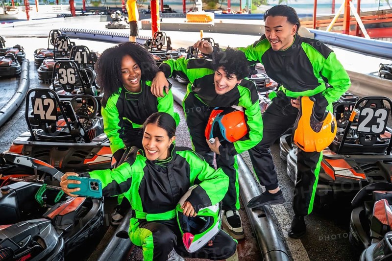 Diverse group of friends in racing suits and helmets smiling for a selfie at a karting track, enjoying an energetic outdoor motorsports outing and capturing a joyful memory