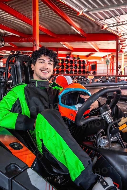 Young man in green and black racing suit smiles holding orange and blue helmet while seated in a go kart on an indoor track, ready for fast, fun karting action