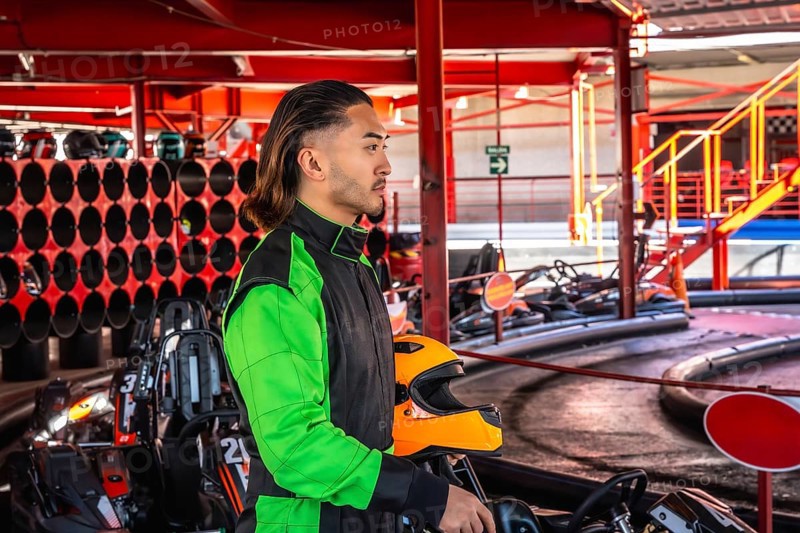 Young man wearing a racing suit standing with an orange helmet by a go kart, preparing to drive on a karting circuit, showcasing leisure sport and adrenaline