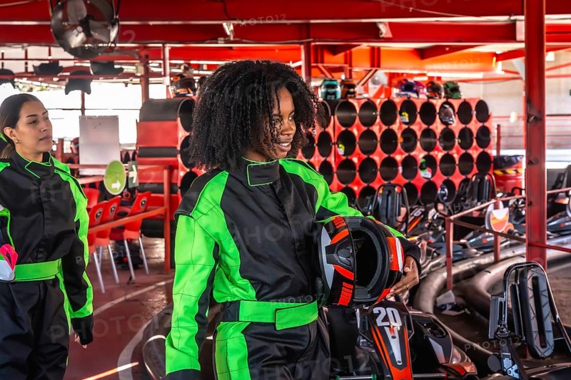 Two cheerful young women in racing suits and holding helmets preparing to race go karts at an indoor track, enjoying a fun day of speed and competition