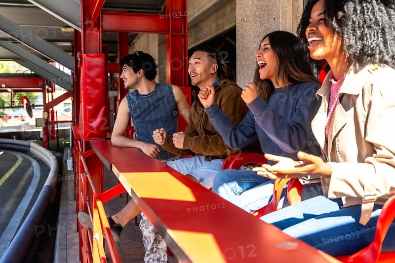 Diverse group of young adult friends are enthusiastically watching and cheering a karting race from the sidelines, expressing excitement and support for the participants
