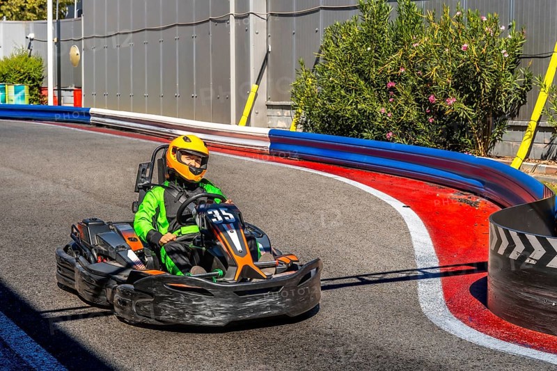 Person wearing a green racing suit and a yellow helmet is driving a go kart around a curve on an outdoor racing track, enjoying the speed and competition involved in motorsport recreation