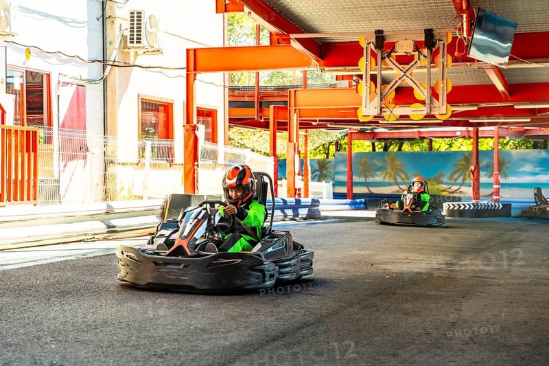 People wearing helmets and green racing suits driving go karts around an outdoor track, enjoying the thrill of speed and competition during a sunny day