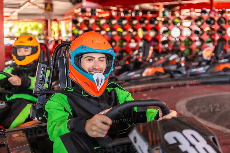 Young man wearing a colorful helmet and racing suit, smiling and driving a go kart on a track with other karts and a helmet wall in the background, conveying excitement and fun